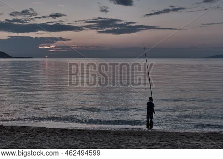Silhouette Of Boy Fishing With Fishing Rod On Sandy Shore Of Bay. Summer Landscape Of Nature. Sunset