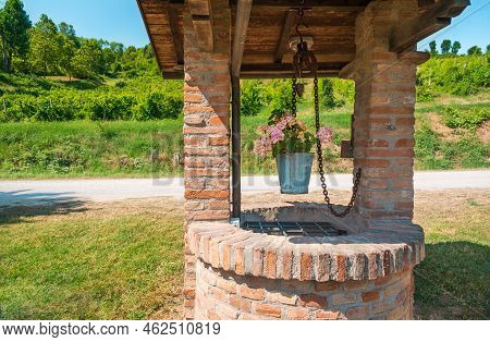 A Brick Well Stands By The Road Near Green Hills With A Hanging Bucket In Which A Pink Flower Blooms