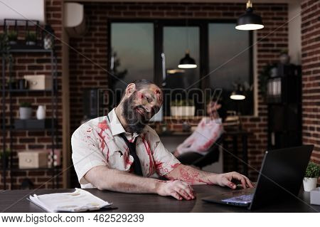 Portrait Of Zombie Businessman Sitting At Desk, Looking Creepy And Terrifiyng In Company Office. Bra