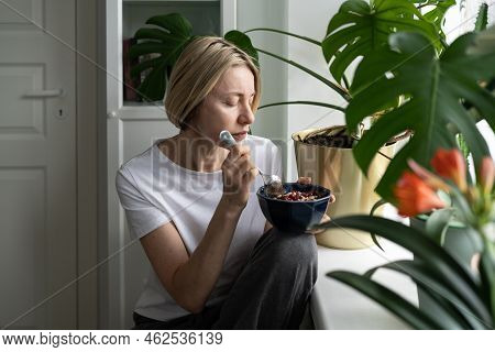 Tranquil Mature Woman Eats Breakfast In Bowl Sitting Alone Near Windowsill With Blooming Houseplant.