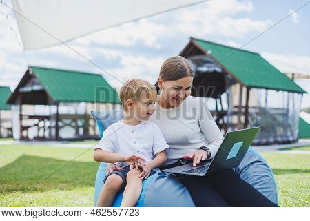 A Mother With A Laptop, Her Son Sitting Next To Her Outdoors On A Green Grass Lawn. Rest And Work Wi