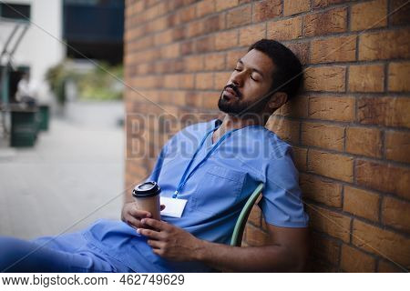 Tired Caregiver Man Resting Outdoor, In Front Of Nurishing Home.