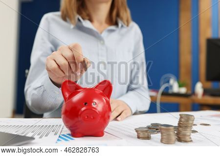A Woman In The Office Puts A Coin In A Red Piggy Bank, Close-up. Capitalization Of Income, Saving Mo