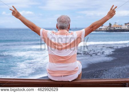 Rear View Of Senior Man Sitting On A Bench Face To The Sea Gesturing Victory With Hands Looking At H