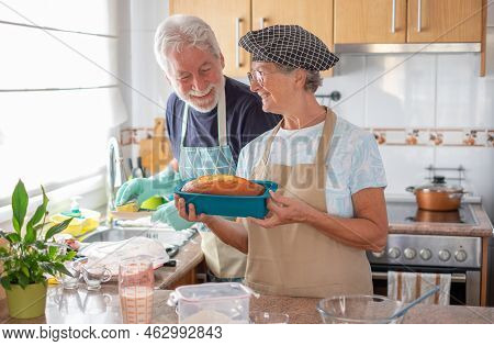 Smiling Older Woman Showing Her Freshly Baked Homemade Plumcake While Her Husband Washes The Dishes.