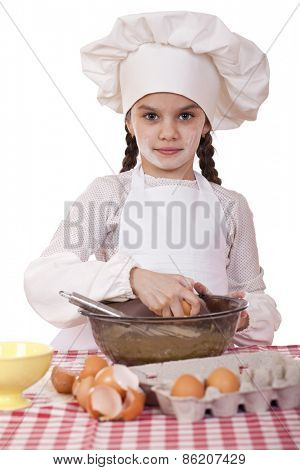 Little cook girl in a white apron breaks eggs in a deep dish, isolated on white background