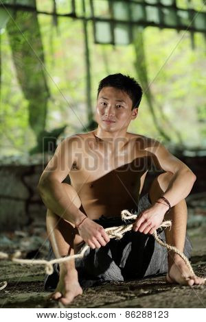 young man in ragged pants sits holding rope in his hands in abandoned building 