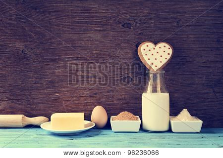 a heart-shaped cookie and the ingredients for cook it, such as milk, eggs, flour, butter and sugar on a blue rustic wooden surface, with a filter effect