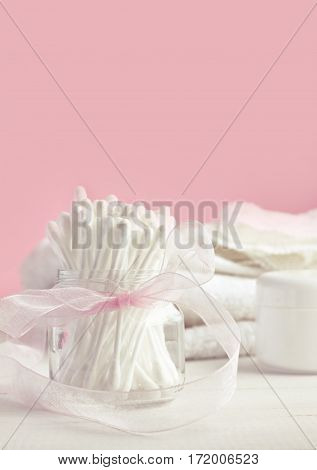 White cotton swabs in glass jar with ribbon bow decor, pink background. Feminine beauty care. Vertical set. Soft light and focus.