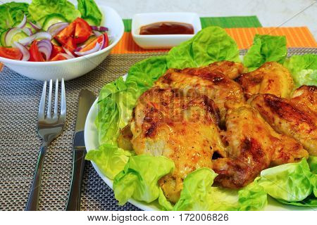 Grill wings on lettuce leaves. Vegetable salad in a white salad bowl. Ketchup in a gravy boat. Table setting.