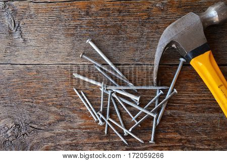 A top view image of an old used yellow hammer and a pile of nails on a wooden workbench.
