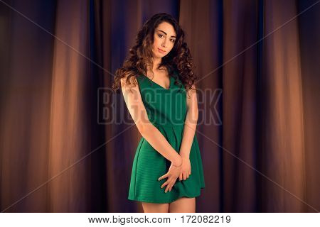 Fashion studio shot of beautiful brunette woman with makeup and curly hairstyle wearing evening green dress on brown silk background.