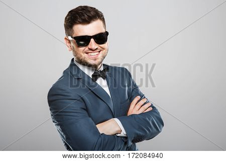 Stylish young man in suit with bow and sunglasses. Hands crossed, looking at camera and smiling. Jacket, bow, official outlook. Waist up, studio, indoors