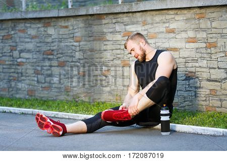 Pain in leg, sport injury. Man sitting and holding his leg. Sportsman limbering up leg, bottle with water standing next to him. Full body, outdoors