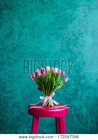 Bouquet of pink and white tulips with pink ribbon on the green painted wall background standing on pink chair. Empty space for text.
