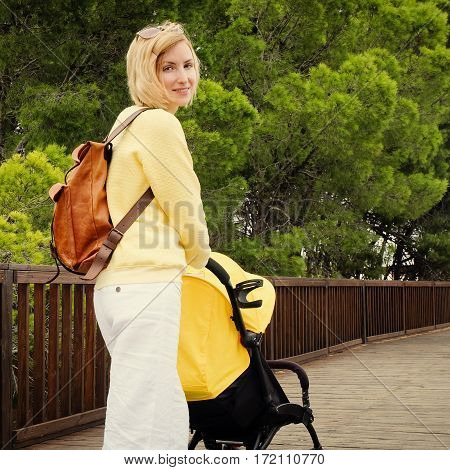 Smiling young mother strolling with newborn in carriage. Blonde hair woman walking in park with stroller