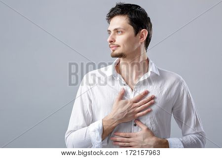 handsome young proud man in a white shirt holding hand on chest on grey background.