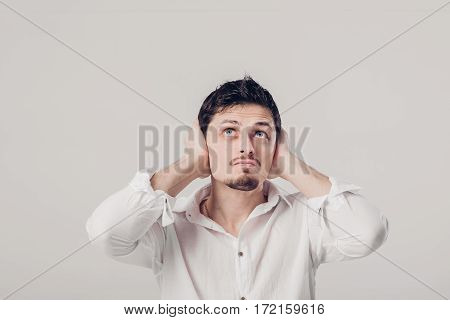 portrait of a young Brunet man in white shirt covers his ears with his hands against a grey background. I don't want to hear anything. soft light