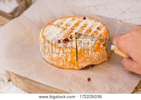 Woman's hand cutting whole wheel of soft French German cheese with orange rind with mold on parchment paper wood cutting board rustic kitchen interior
