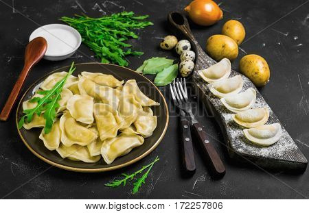 Dumplings with potatoes in plate on black background. On board cooking raw dumplings with potatoes. Ingredients for dumplings with potatoes onions arugula eggs bay leaf potatoes sour cream sauce.