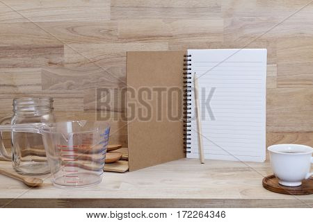 blank notebook with coffice cup and measuring cup and wooden spoon cooking supplies on wood background.