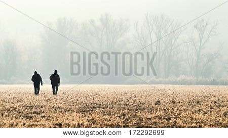 Panorama with silhouette of the two hunters photographer hikers or tourists on the field at sunrise. Group of people going up in the early morning in a rural field through the grass during hunting. Dreamy fogy landscape spring orange misty sunrise