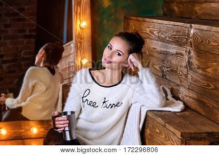 Young brunette holdind a thermocup and sitting in front of mirror with lights