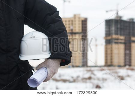 Architect or engineer holding yellow helmet for workers security on background of new highrise apartment buildings and construction cranes on background of winter landscape. Crane lifts load