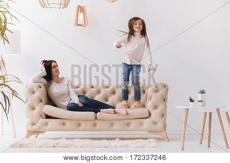 Time with family. Pleasant joyful attractive woman lying on the sofa and smiling while watching her daughter jumping