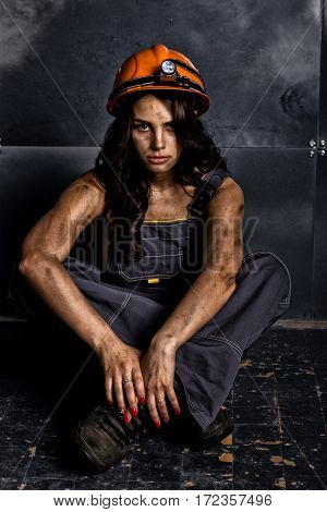 sexy female miner worker with pickaxe, in coveralls over his naked body, sitting on the floor on backdrop of steel wall.