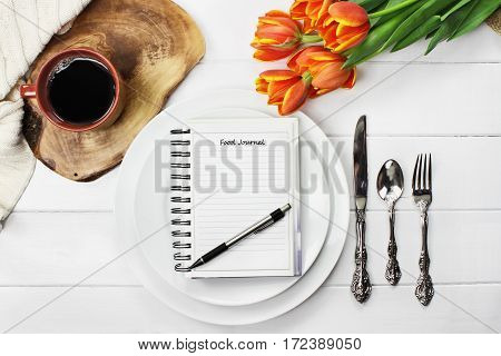 Overhead shot of a food journal over empty plates with a cup of coffee and a bouquet of springtime tulip flowers over white wood table top. Flat lay top view style.