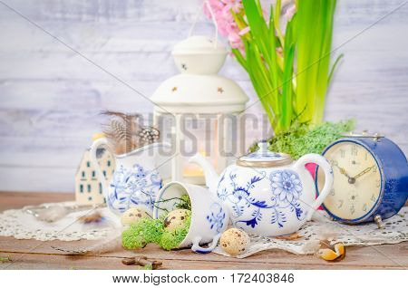 Spring floral still life with hyacinth. Blue alarm clock, teapot, cup, moss. Lantern, feathers and quail eggs on wooden background