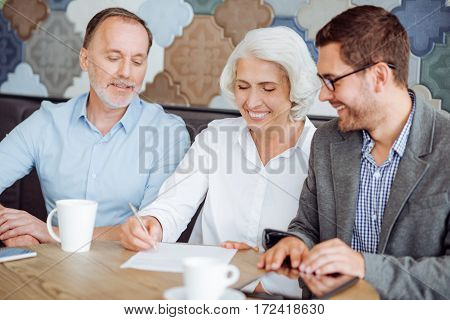 Put your signature. Cheerful delighted senior couple sititng at the table and signing the contract while meeting with insurance manager
