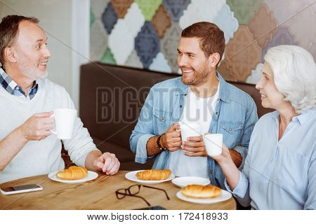 Family meeting. Positive smiling aged couple and their grandson sitting at the table and having lunch while resting in the cafe