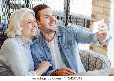 Just smile. Cheerful content smiling man and his grandfather resting in the cafe while makign selfies
