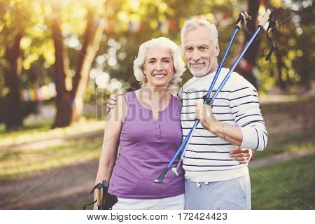 Active way of life. Positive delighted elderly man standing near his wife and hugging her while holding walking poles