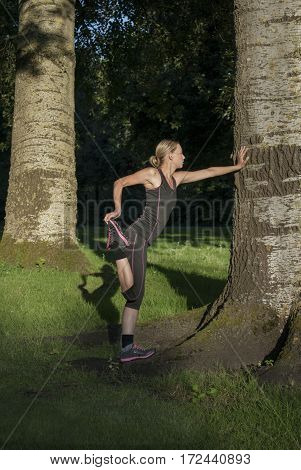 Beautiful young woman doing stretching exercises for quadriceps in park during sundown in summer