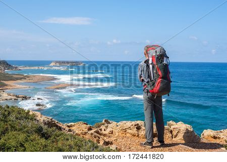Traveler with backpack standing on the rocky sea shore and watching the surf. Sunny day on the peninsula Akamas Cyprus. Back view.