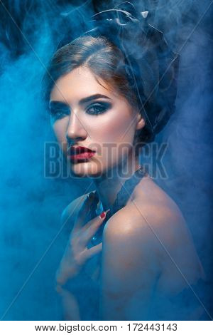Beautiful young woman with bright makeup, red lips, black shawl on head and necklace. Studio beauty shot with blue smoke. Copy space.