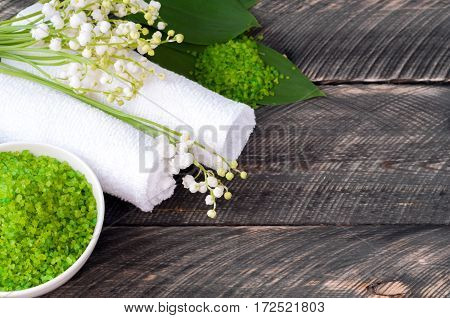 Natural green sea salt and towel for bath and spa on a wooden board. Lilies of the valley in the background. Selective focus