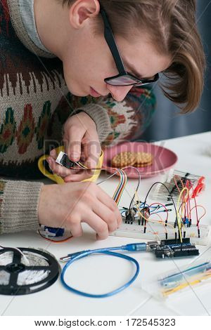 Young geek connecting wires to breadboard. Attractive student in glasses and old pulover invent new gadget at university laboratory. Electronic construction, development, diy, hobby concept