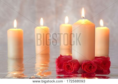Group of burning candles with reflection and a group of red roses surrounding the candle in the foreground