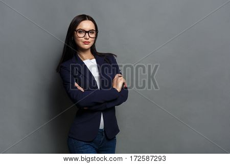 Beautiful young caucasian businesswoman studio shot on grey background. Charming and confident serious brunette woman in casual dark blue jacket wearing glasses