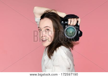 Portrait of the girl with camera in her right hand and her left hand touching her brown hair on the pink background