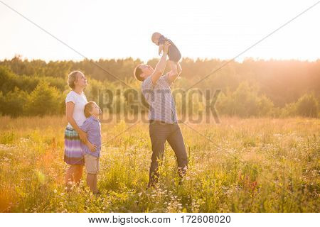 Happy family with two boys on a summer meadow enjoying sunset. Mother and father with kid boy and cute baby. Family together parents with their little children. Father raising baby up in the air.