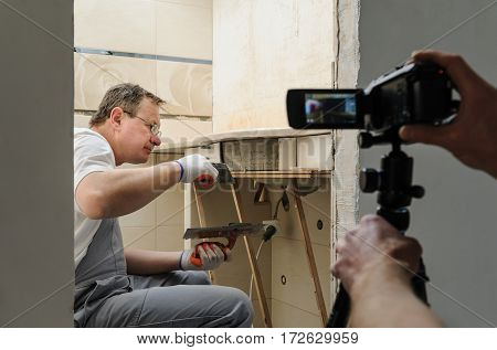 Worker installs ceramic tiles. The operator is shooting this process using camcorder and tripod.