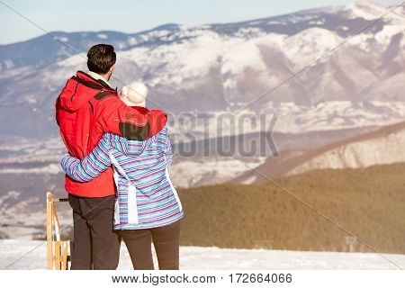 Rear view of a loving couple in fur hood jackets looking at snowed mountain range.