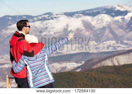 Rear view of a loving couple in fur hood jackets looking at snowed mountain range.