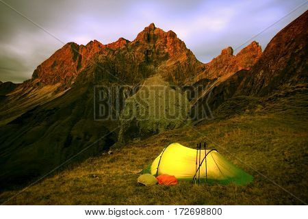 Last glow of daylight hits the mountain peaks. The warm and illuminated tent promises a comfortable night.