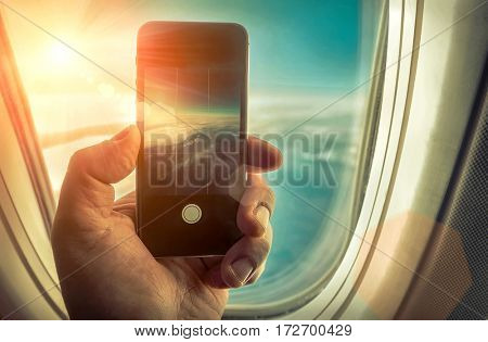 Hand with cameraphone shooting sky with clouds under sun light in airplane. View from porthole at sunny day.